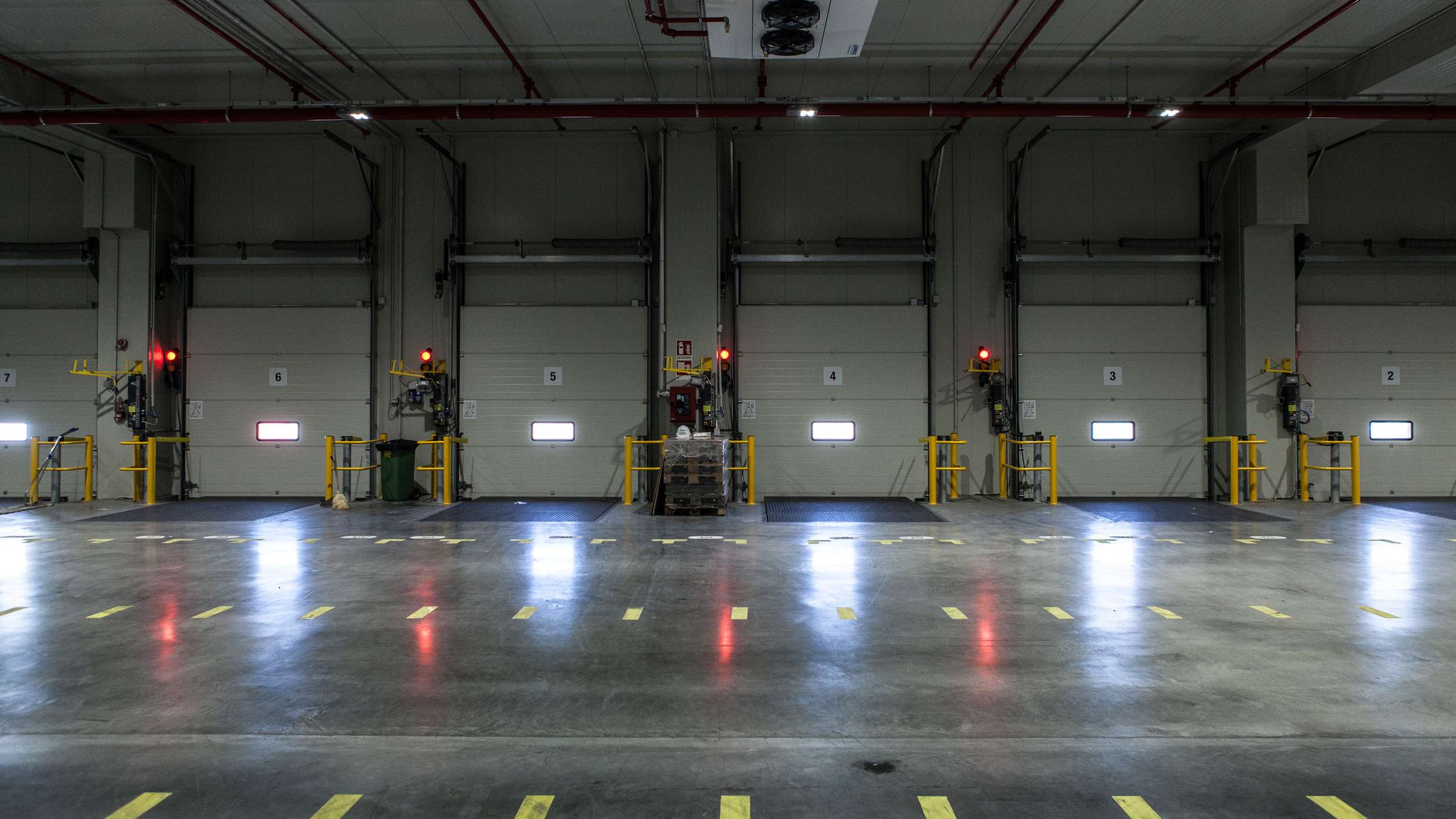 In a gloomy warehouse, the loading ramps can be seen from inside. Red spotlights and a little light from outside illuminate the room.