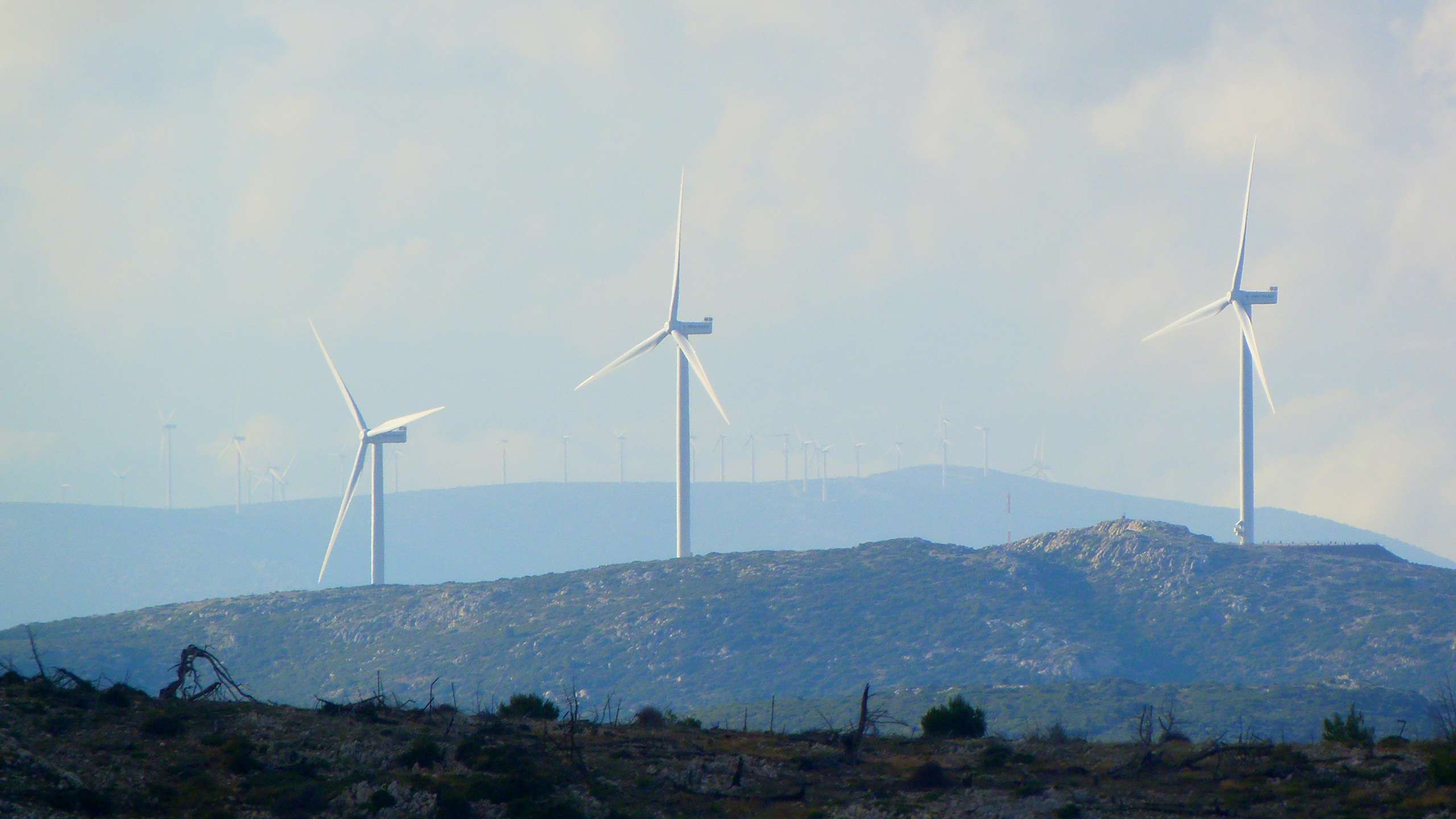 Tre turbine eoliche si ergono su una collina. Il cielo è nuvoloso e colora il paesaggio di blu e grigio.