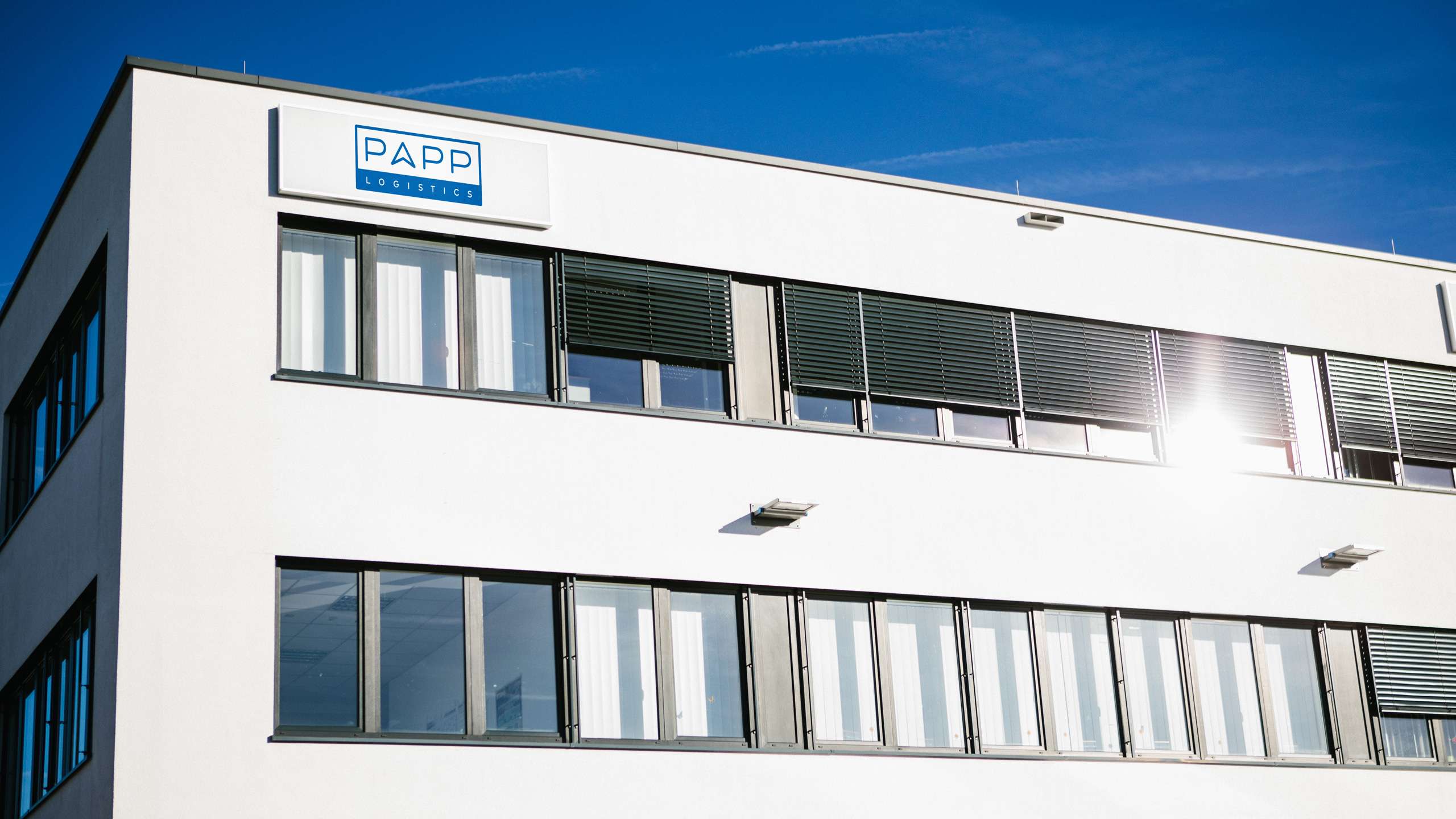 Ein weißes Gebäude vor einem blauen, wolkenlosen Himmel. Am oberen linken Eck steht das PAPP-Logo. Zwei Fensterreihen werden von der Sonne beschienen.