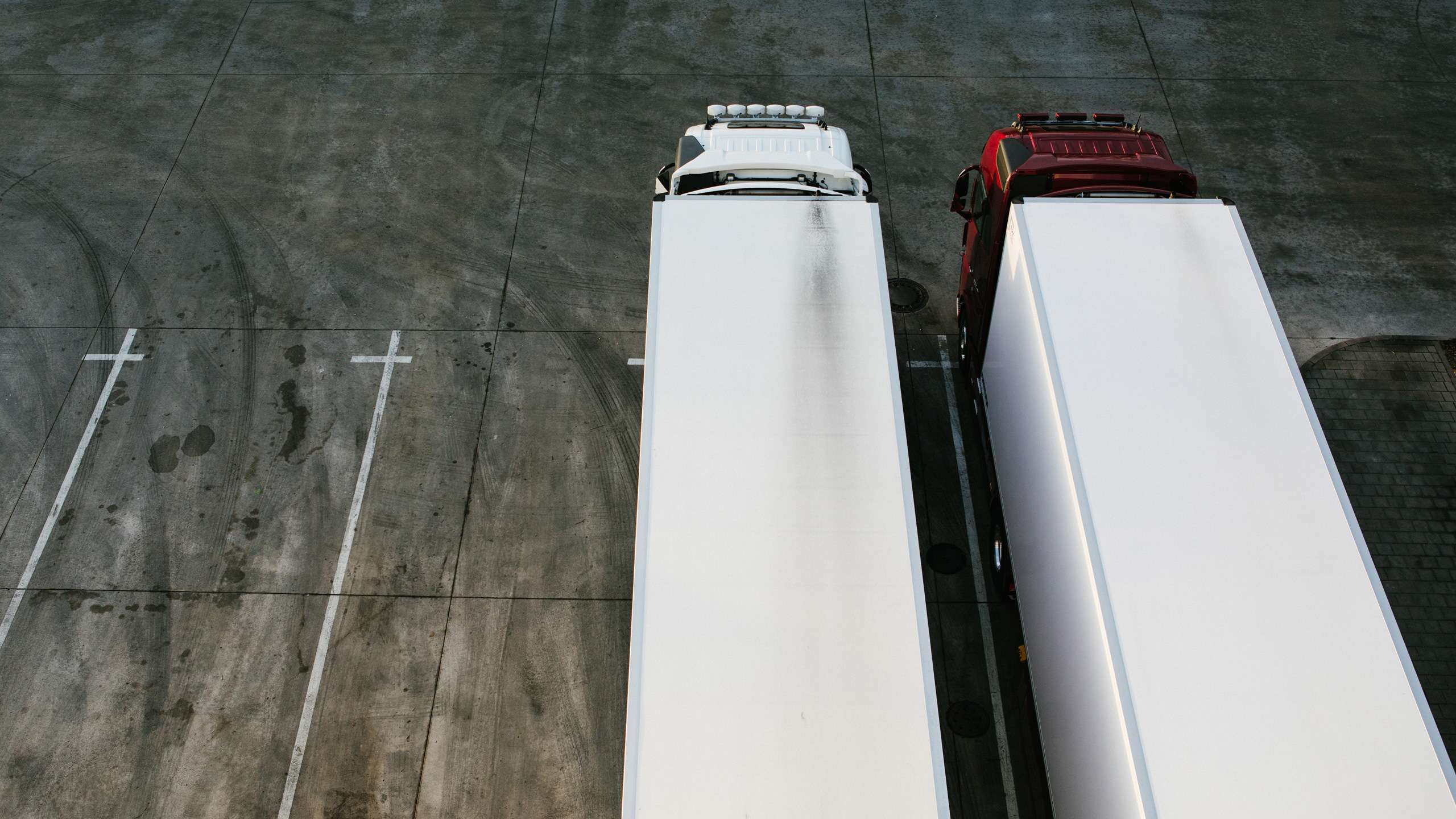 Two trucks are standing next to each other in a truck park, seen from above. The one on the left is completely white, the one on the the right has a shiny red cab.