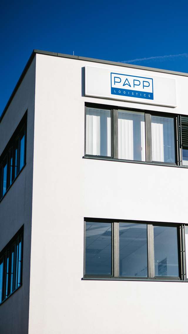 Ein weißes Gebäude vor einem blauen, wolkenlosen Himmel. Am oberen linken Eck steht das PAPP-Logo. Zwei Fensterreihen werden von der Sonne beschienen.