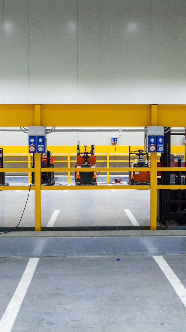 Four red pallet trucks are parked next to each other in the warehouse. A yellow railing runs in the foreground.