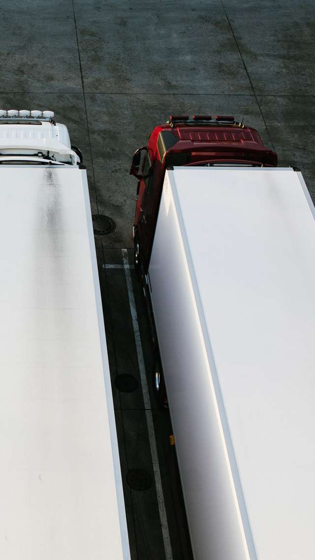 Two trucks are standing next to each other in a truck park, seen from above. The one on the left is completely white, the one on the the right has a shiny red cab.