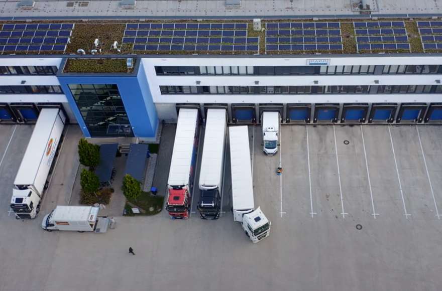 Da una visuale aerea si vedono i camion fermi sulle rampe di carico davanti a un magazzino. Sulla sinistra si vede un vano scale blu.