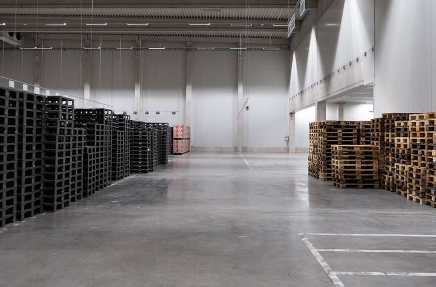 In a warehouse with a grey concrete floor, there are stacked plastic pallets on the left and wooden Euro pallets on the right.