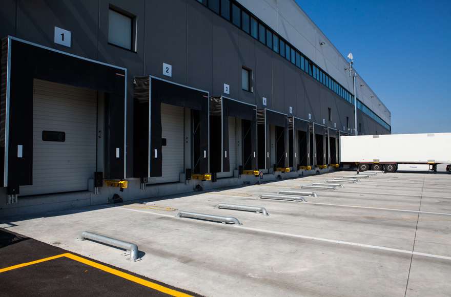 The loading ramps of a dark grey warehouse are seen against a blue sky. In the background, a truck docks at a loading ramp.