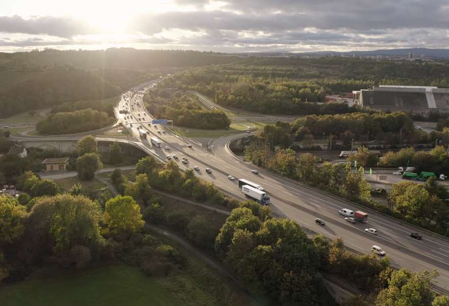 A bird's eye view of a stretch of motorway on which a PAPP truck is driving through a green landscape. Evening sunlight breaks through a cloudy sky.