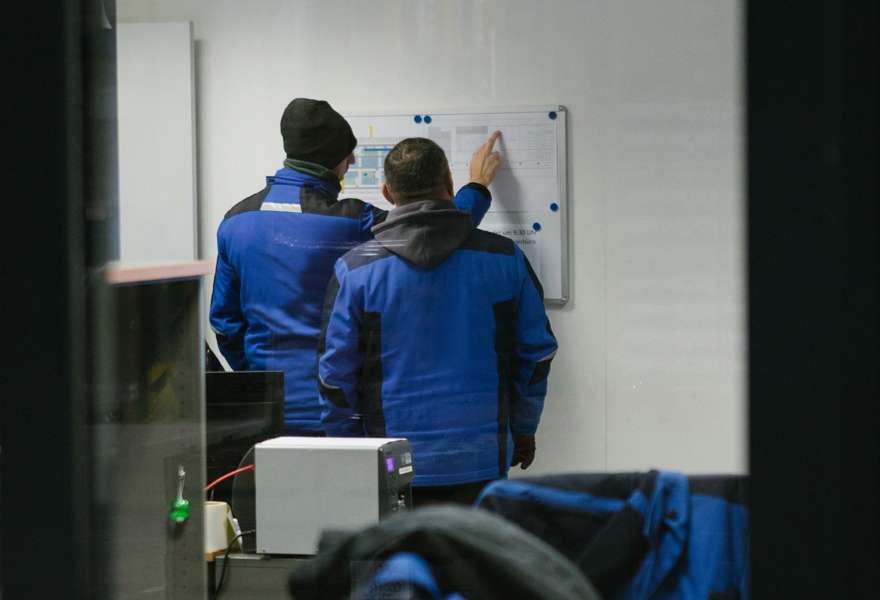 Two PAPP employees in blue jackets stand behind a window in front of a magnetic board and point to pinned documents.