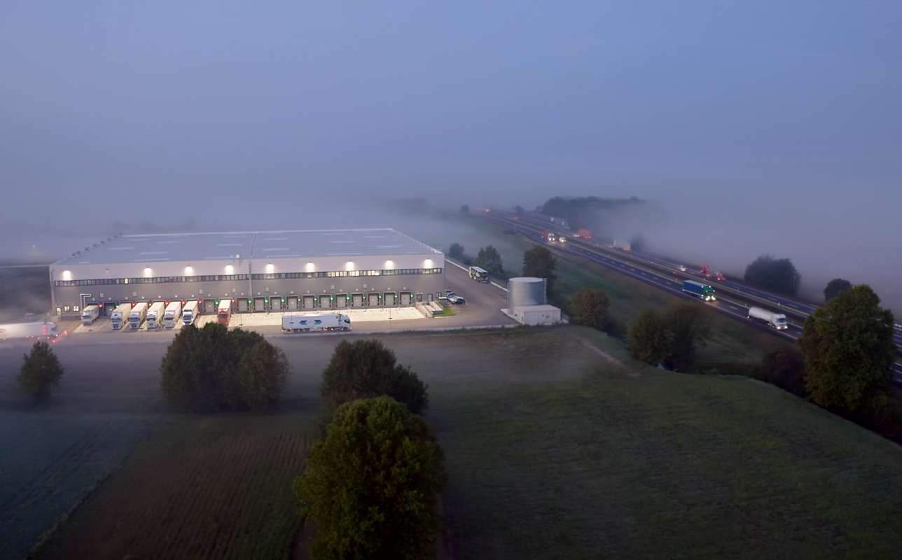 Eine PAPP Lagerhalle liegt früh am Morgen hell erleuchtet am Rande einer Autobahn. LKWs stehen an den Laderampen und fahren in die vernebelte, grüne Landschaft.