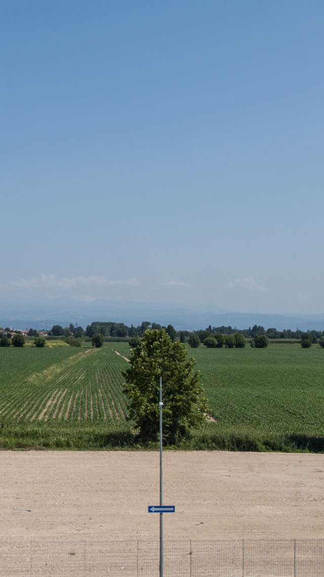 Ein Panoramablick auf grüne Felder und Bäume vor blauem Himmel und fernen Bergen. Im Vordergrund sieht man  Erde und Straßenschildern, die eine Einbahnstraße kennzeichnen.